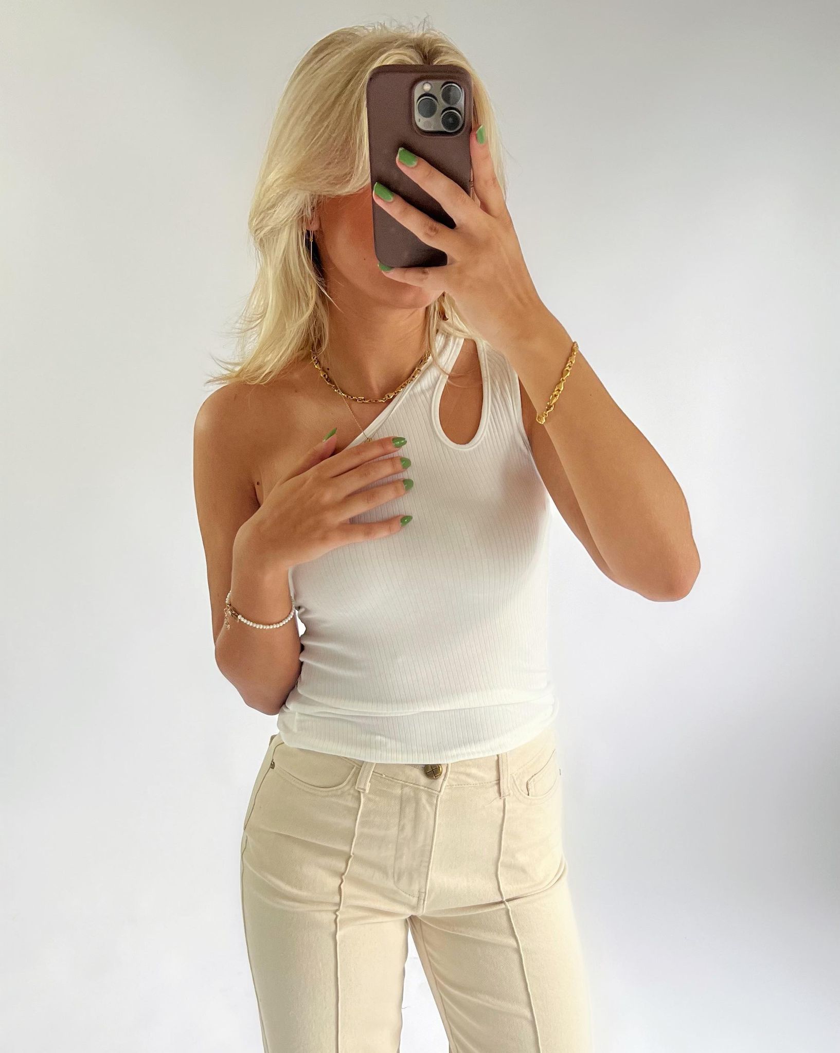 En person klædt i en hvid, skulderfri top tager et spejlfoto med en smartphone. De bærer beige bukser og guldtilbehør. Baggrunden er neutral og lys.