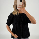 Person tager et spejlbillede iført sort striktrøje og grå jeans, med en smartphone foran ansigtet. Baggrunden er ensfarvet lys.