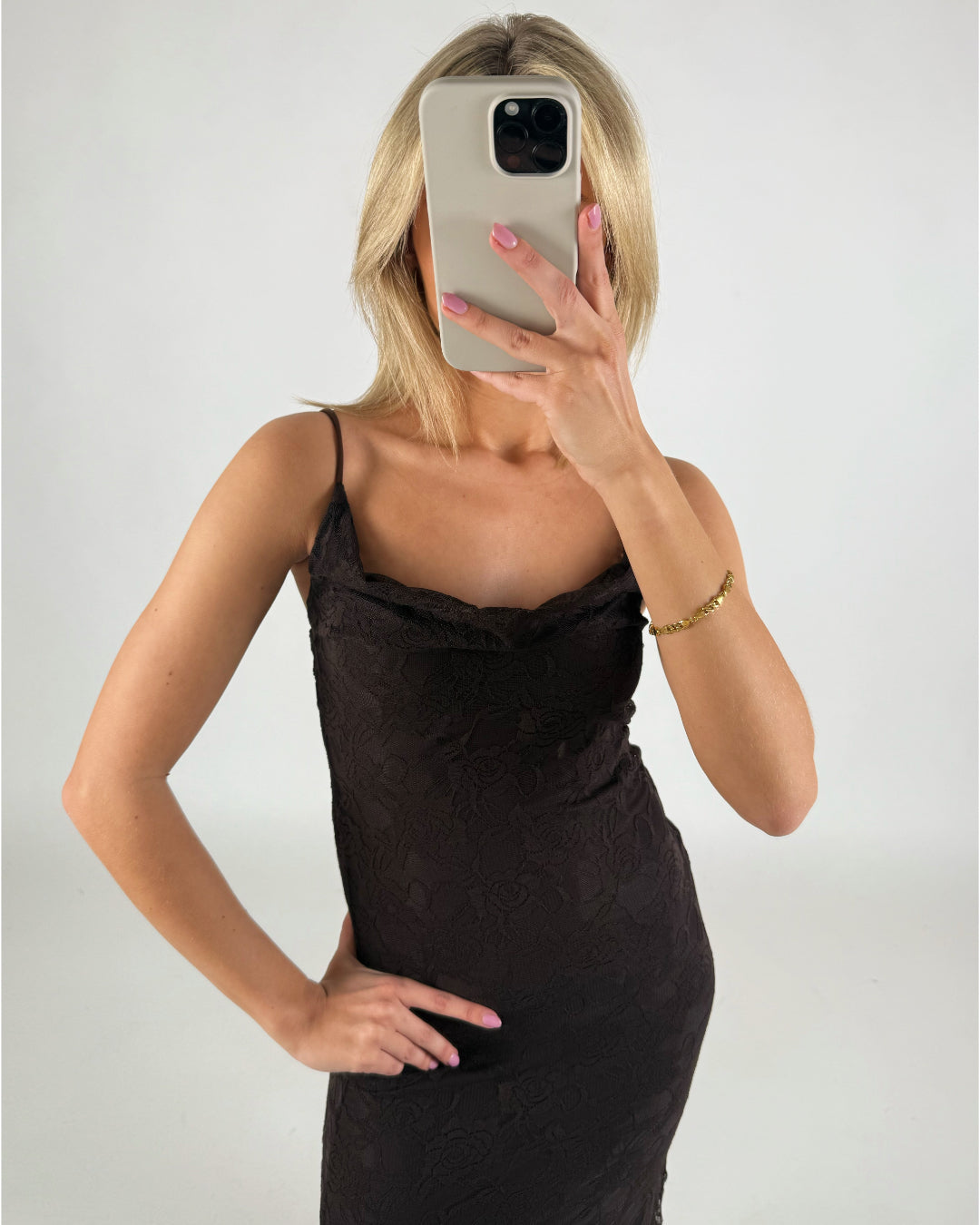 En person i en sort kjole tager en selfie med en smartphone, som dækker ansigtet. Personen står i et lyst rum og bærer et gyldent armbånd.