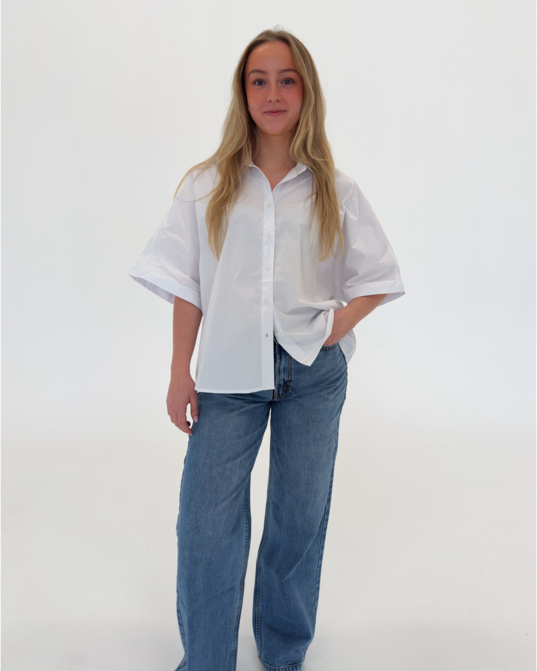 En person står afslappet i en hvid skjorte og blå jeans, med hånden i lommen, på en ensfarvet hvid baggrund.
