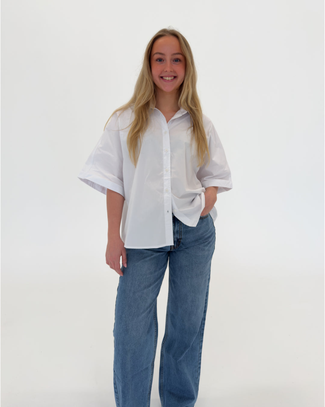 En person i en hvid skjorte står og smiler, med hånden i lommen på jeans. Baggrunden er ensfarvet og neutral, uden tekst.