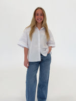 En person i en hvid skjorte står og smiler, med hånden i lommen på jeans. Baggrunden er ensfarvet og neutral, uden tekst.