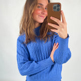 En person iført en blå sweater tager en selfie med en mobiltelefon. Personen smiler, og baggrunden er neutral med en hvid baggrund. Ingen tekst er til stede.