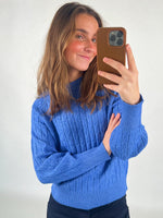 En person iført en blå sweater tager en selfie med en mobiltelefon. Personen smiler, og baggrunden er neutral med en hvid baggrund. Ingen tekst er til stede.