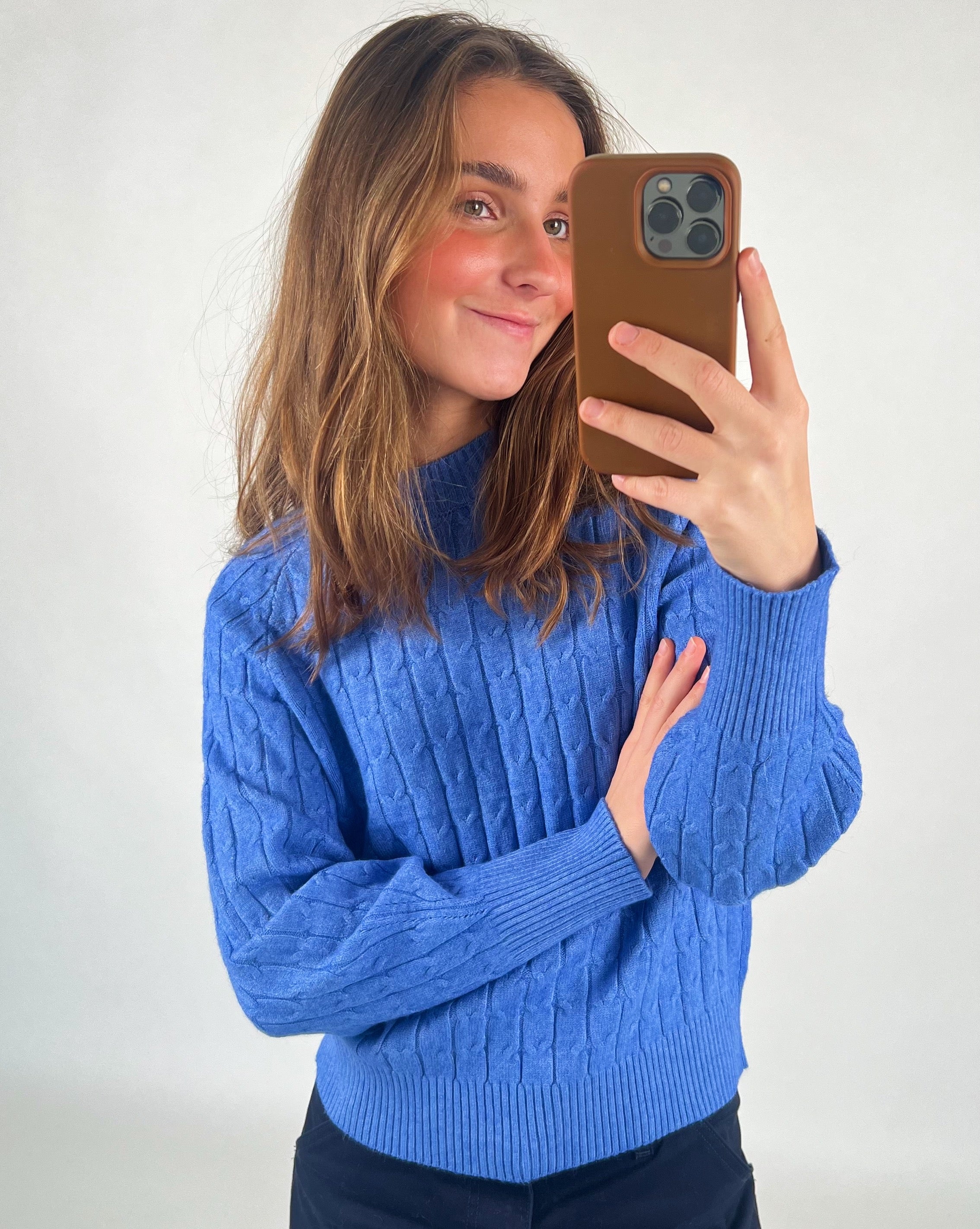 En person iført en blå sweater tager en selfie med en mobiltelefon. Personen smiler, og baggrunden er neutral med en hvid baggrund. Ingen tekst er til stede.