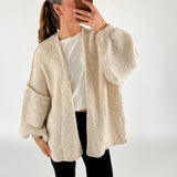 En person tager en selfie iført en hvid trøje og beige strikcardigan, med en neutral baggrund. Personen har en hestehale og bærer sort bukser samt gyldent smykke.