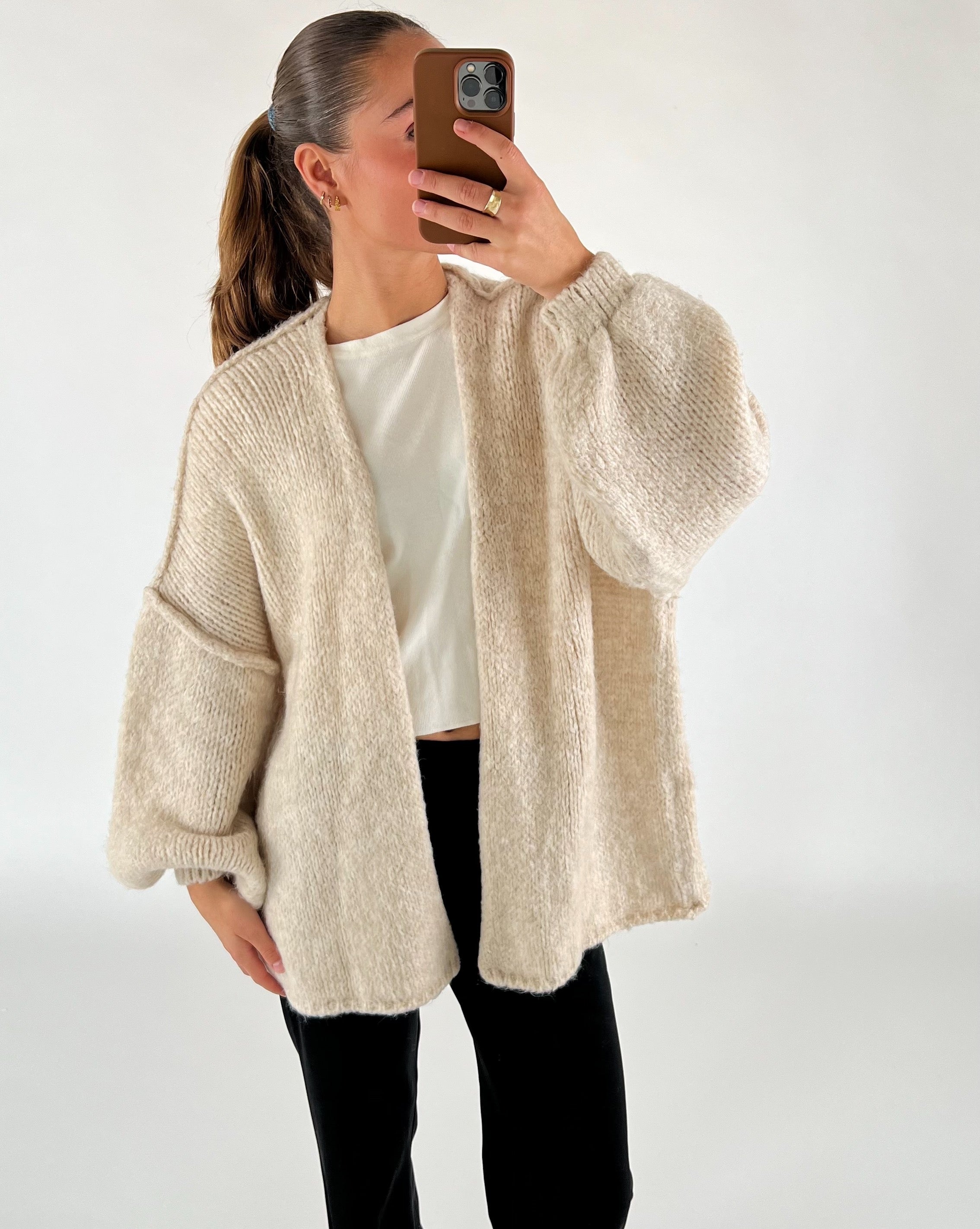 En person tager en selfie iført en hvid trøje og beige strikcardigan, med en neutral baggrund. Personen har en hestehale og bærer sort bukser samt gyldent smykke.