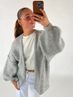 En person tager en selfie iført en grå striktrøje og hvid bluse. Personen holder en telefon med brun cover i et lyst rum.