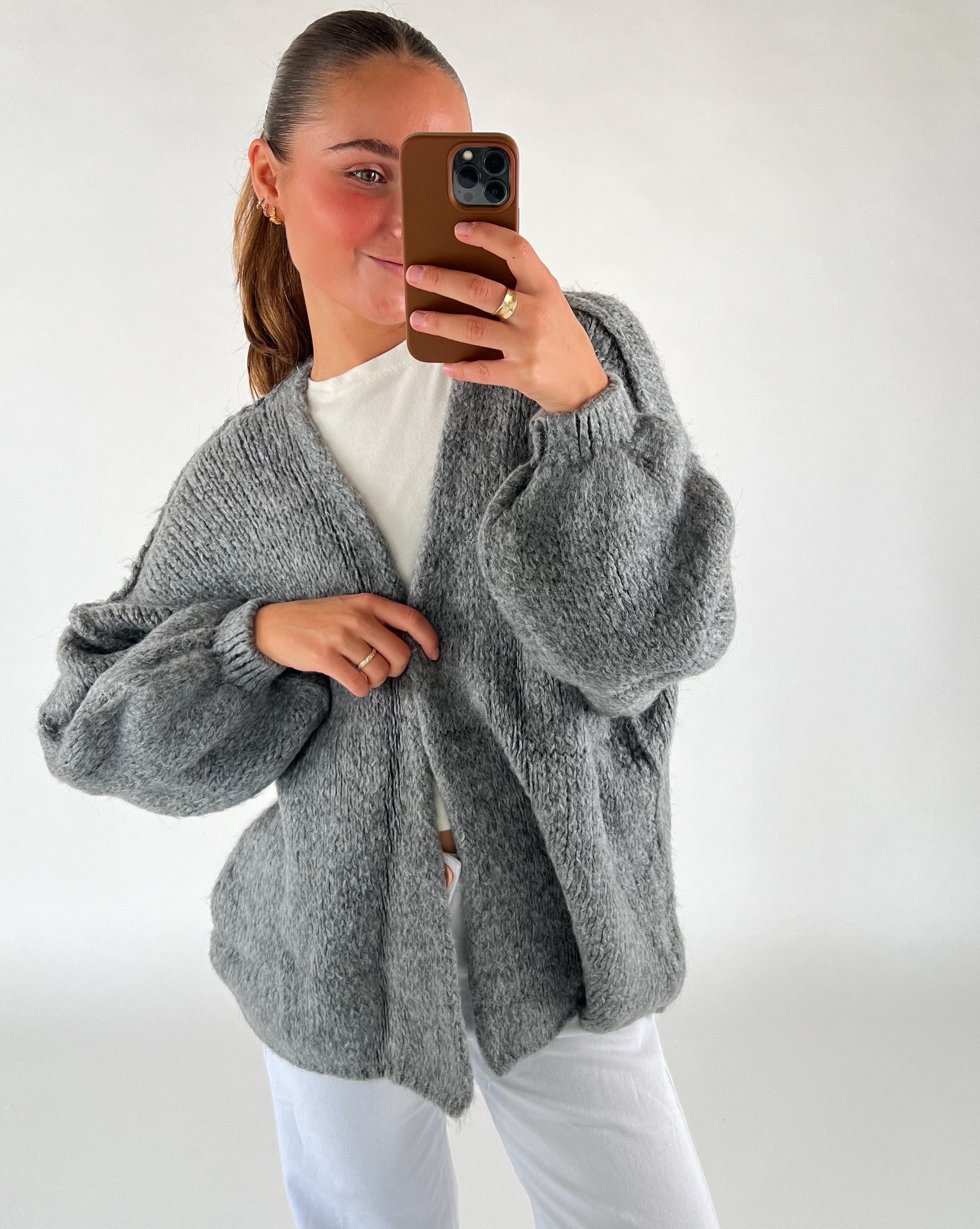 En person iført en grå, strikket cardigan tager en selfie med en telefon i en brun etui. Personen bærer en hvid t-shirt og hvide bukser i et lys rum.