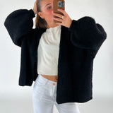En person i sort sweater og hvide bukser tager et spejlfoto med en telefon i et minimalistisk rum. Personen holder håret tilbage med en hånd.