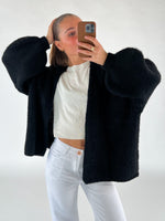 En person i sort sweater og hvide bukser tager et spejlfoto med en telefon i et minimalistisk rum. Personen holder håret tilbage med en hånd.
