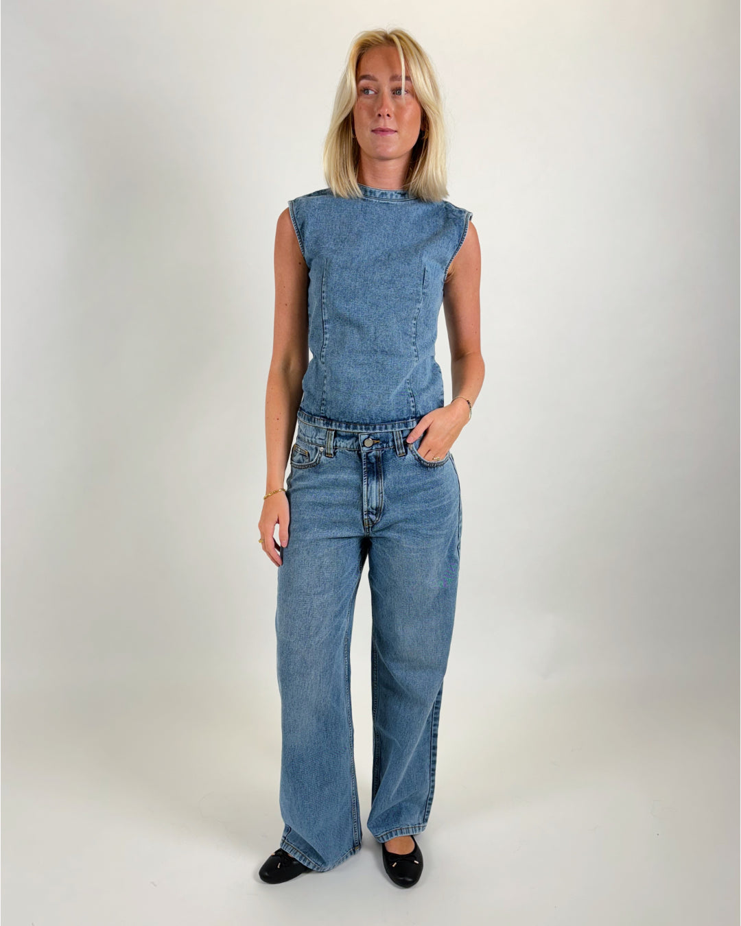 En person iført en ærmeløs denim top og jeans står oprejst i et lyst rum, ser fremad. Vedkommende har lyst hår og sorte flade sko.