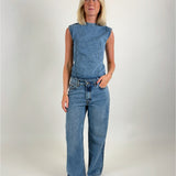 En person iført en ærmeløs denim top og jeans står oprejst i et lyst rum, ser fremad. Vedkommende har lyst hår og sorte flade sko.