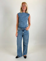 En person iført en ærmeløs denim top og jeans står oprejst i et lyst rum, ser fremad. Vedkommende har lyst hår og sorte flade sko.