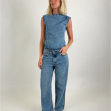 Kvinde iført ærmeløs denimtop og jeans, står med hånden i lommen i et minimalistisk, lyst studie, iført sorte sko.