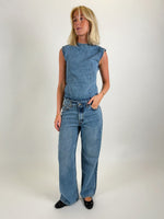 Kvinde iført ærmeløs denimtop og jeans, står med hånden i lommen i et minimalistisk, lyst studie, iført sorte sko.