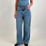 En person bærer en denim jumpsuit og sorte sko, står med en hånd i lommen i et neutralt, lyst miljø.