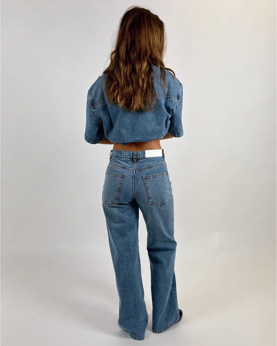 En person med langt brunt hår står med ryggen til, iklædt blå denimjakke og jeans i et enkelt studie med lys baggrund.