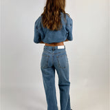 En person med langt brunt hår står med ryggen til, iklædt blå denimjakke og jeans i et enkelt studie med lys baggrund.