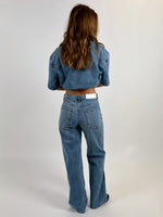 En person med langt brunt hår står med ryggen til, iklædt blå denimjakke og jeans i et enkelt studie med lys baggrund.
