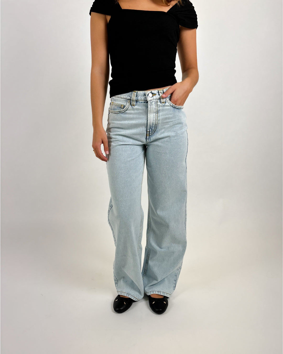 En person iført sort bluse og lyseblå, løstsiddende jeans står med hænderne i lommen. Personen er i et neutralt, lysegråt studie uden yderligere elementer.