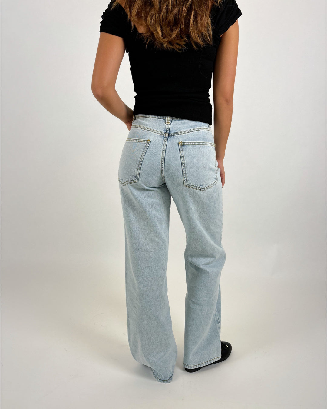 En person iført lys blå jeans og sort t-shirt står med ryggen til i et lyst studiosetup. Personens hår er langt og bølget.