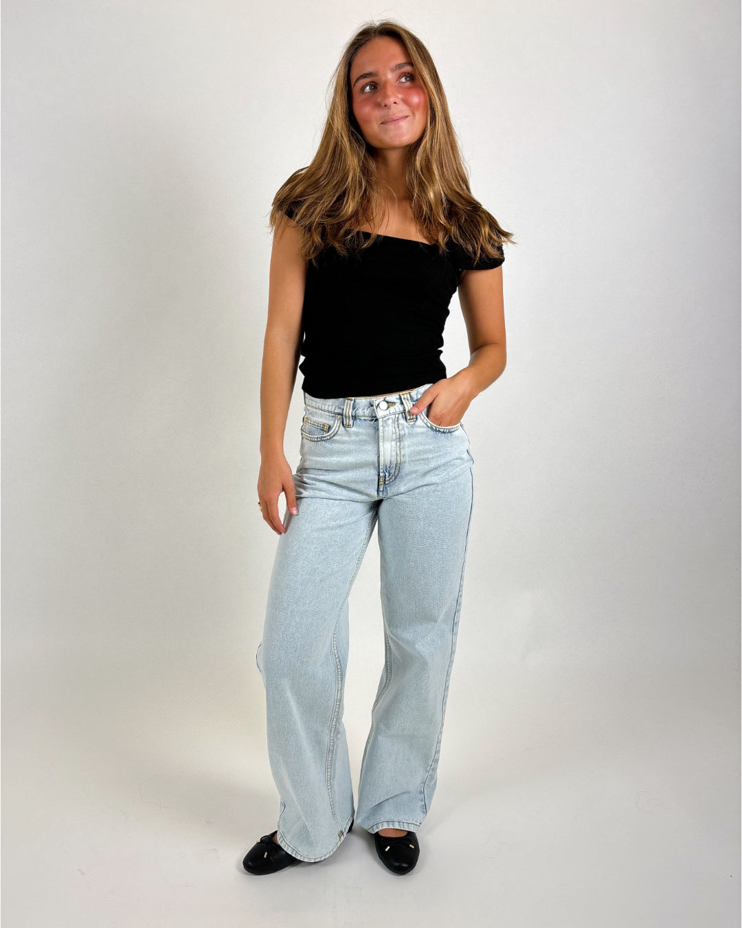 En kvinde står med en hånd i lommen iført en sort top og lyseblå jeans. Baggrunden er en ensfarvet, lys flade.