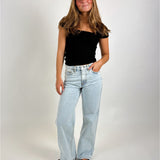En kvinde står med en hånd i lommen iført en sort top og lyseblå jeans. Baggrunden er en ensfarvet, lys flade.