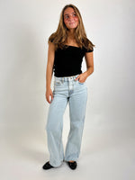 En kvinde står med en hånd i lommen iført en sort top og lyseblå jeans. Baggrunden er en ensfarvet, lys flade.