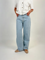 En person står iført lyseblå jeans og en hvid skjorte med korte ærmer og knapper. Personen har hænderne i lommerne i et neutralt miljø. Ingen tekst er til stede.