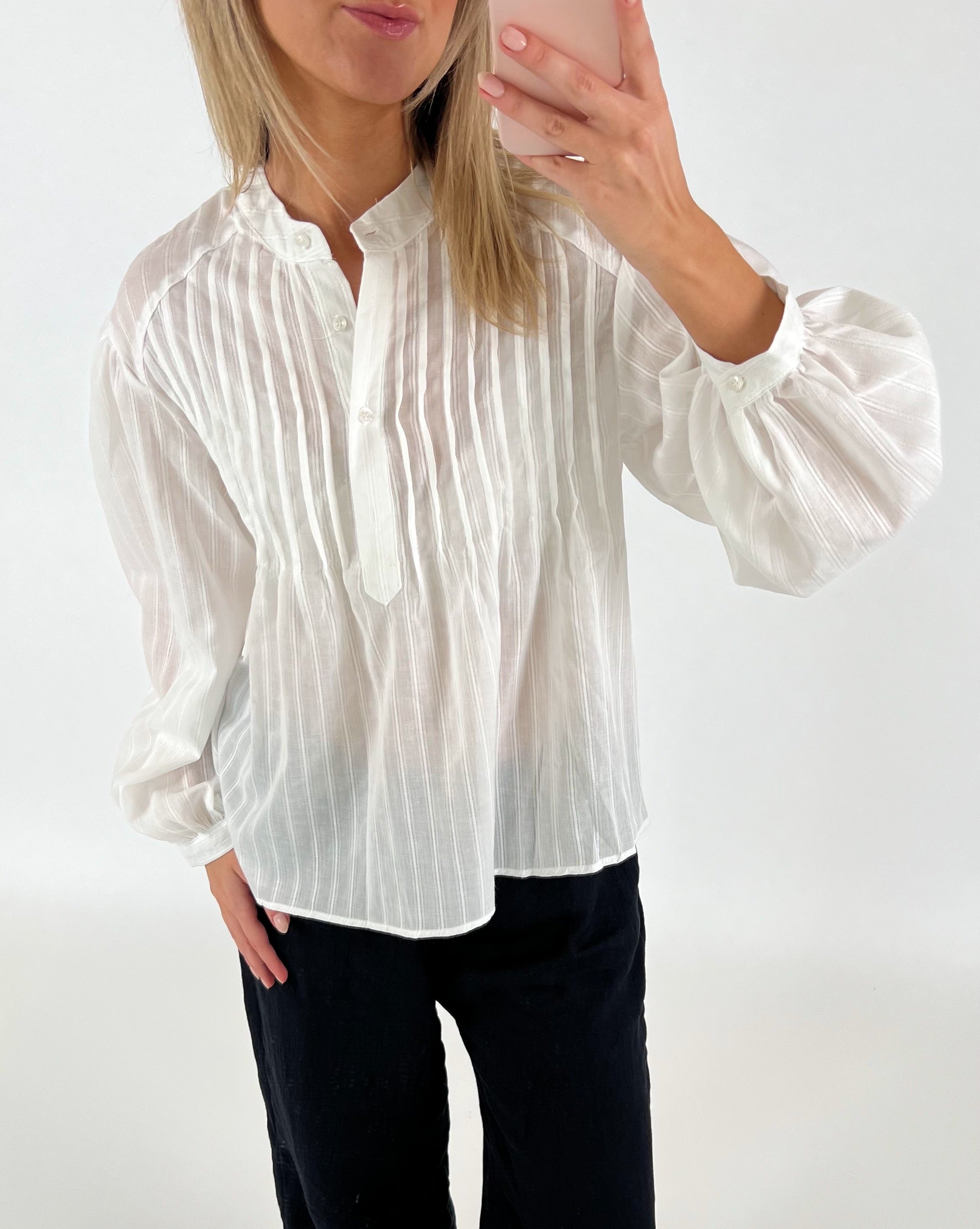 En kvinde iført en hvid, stribet bluse tager et billede med en smartphone. Hun står i et lyst rum, iført sorte bukser.