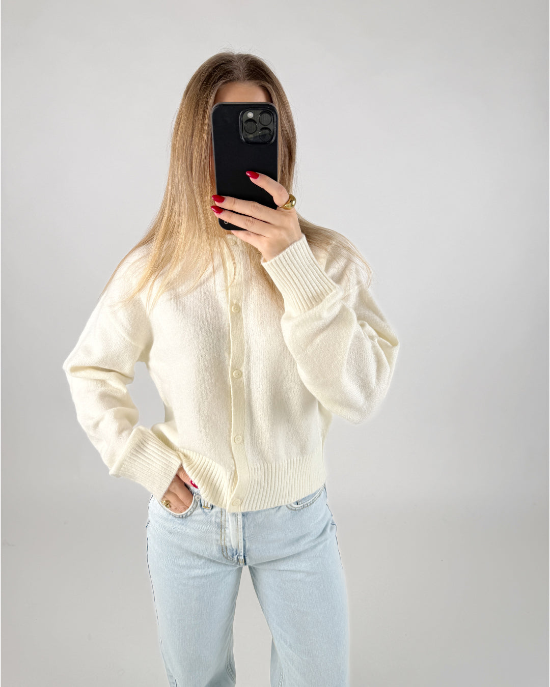 En person i en hvid sweater tager et billede med en mobiltelefon, der dækker ansigtet. De har lyseblå jeans på og står i et minimalistisk rum.