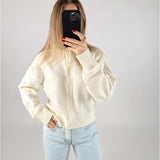 En person i en hvid sweater tager et billede med en mobiltelefon, der dækker ansigtet. De har lyseblå jeans på og står i et minimalistisk rum.