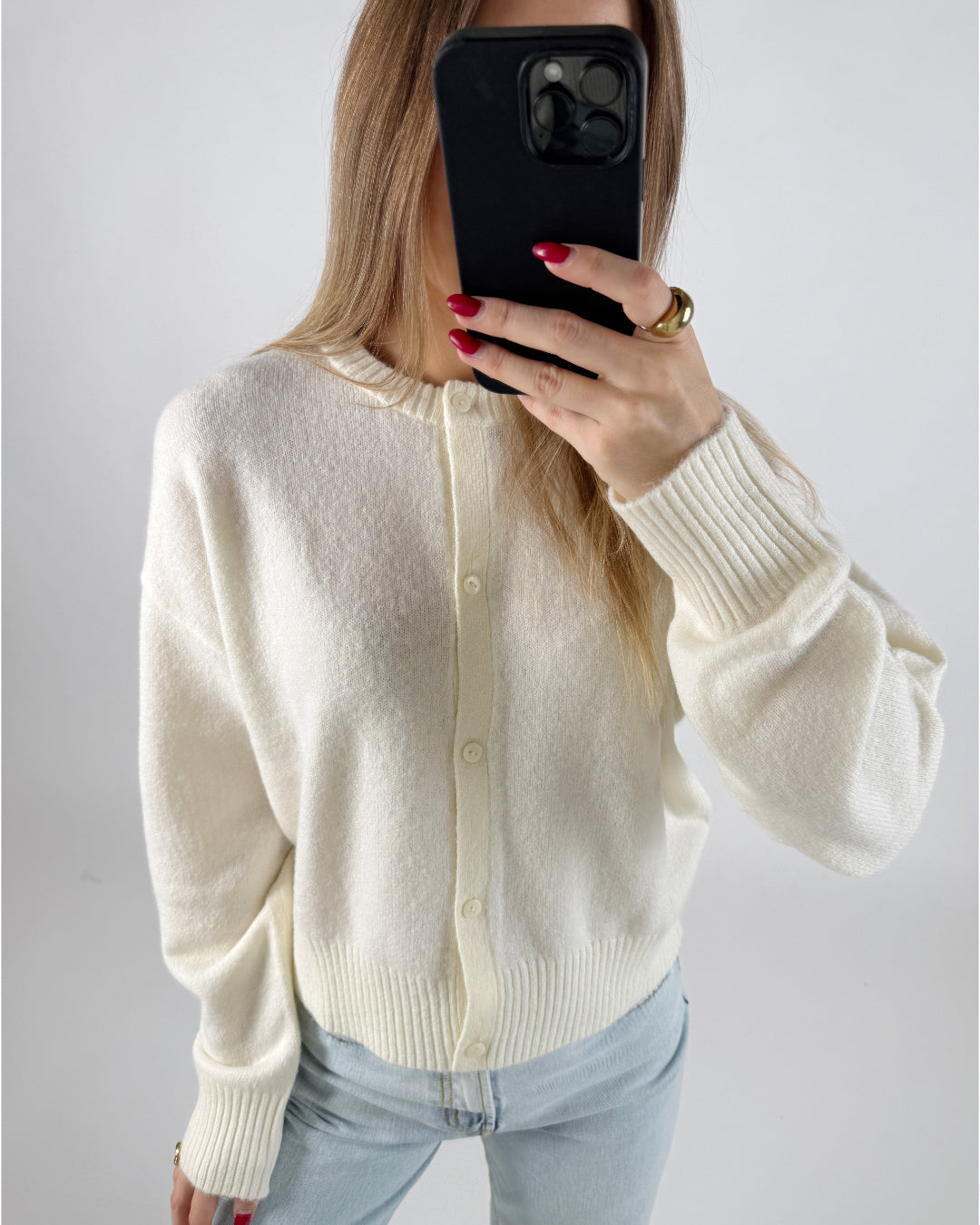 En person holder en telefon foran ansigtet, iført en cremefarvet cardigan og lyse jeans, i et lyst rum. Røde negle og en stor guldring er synlige.