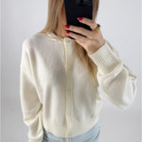 En person holder en telefon foran ansigtet, iført en cremefarvet cardigan og lyse jeans, i et lyst rum. Røde negle og en stor guldring er synlige.