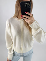 En person holder en telefon foran ansigtet, iført en cremefarvet cardigan og lyse jeans, i et lyst rum. Røde negle og en stor guldring er synlige.