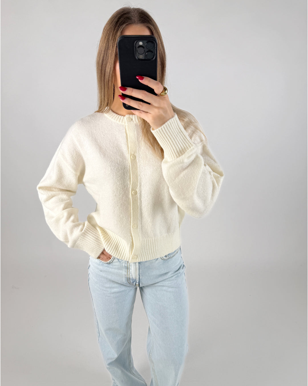 En person iført hvid cardigan og lyseblå jeans tager et spejlbillede med en sort telefon. Personen står i et neutralt belyst rum.