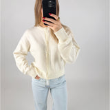 En person iført hvid cardigan og lyseblå jeans tager et spejlbillede med en sort telefon. Personen står i et neutralt belyst rum.
