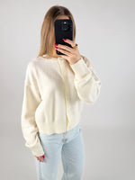En person iført en cremefarvet cardigan tager et spejlbillede med en mobiltelefon. Personen har rødlakerede negle og en stor ring. Baggrunden er hvid og minimalistisk.