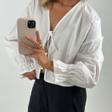 En person iført hvid skjorte tager en selfie med en mobiltelefon med beige cover. De har guldsmykker på og står foran en neutral baggrund.