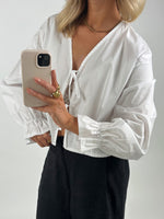 En person iført hvid skjorte tager en selfie med en mobiltelefon med beige cover. De har guldsmykker på og står foran en neutral baggrund.