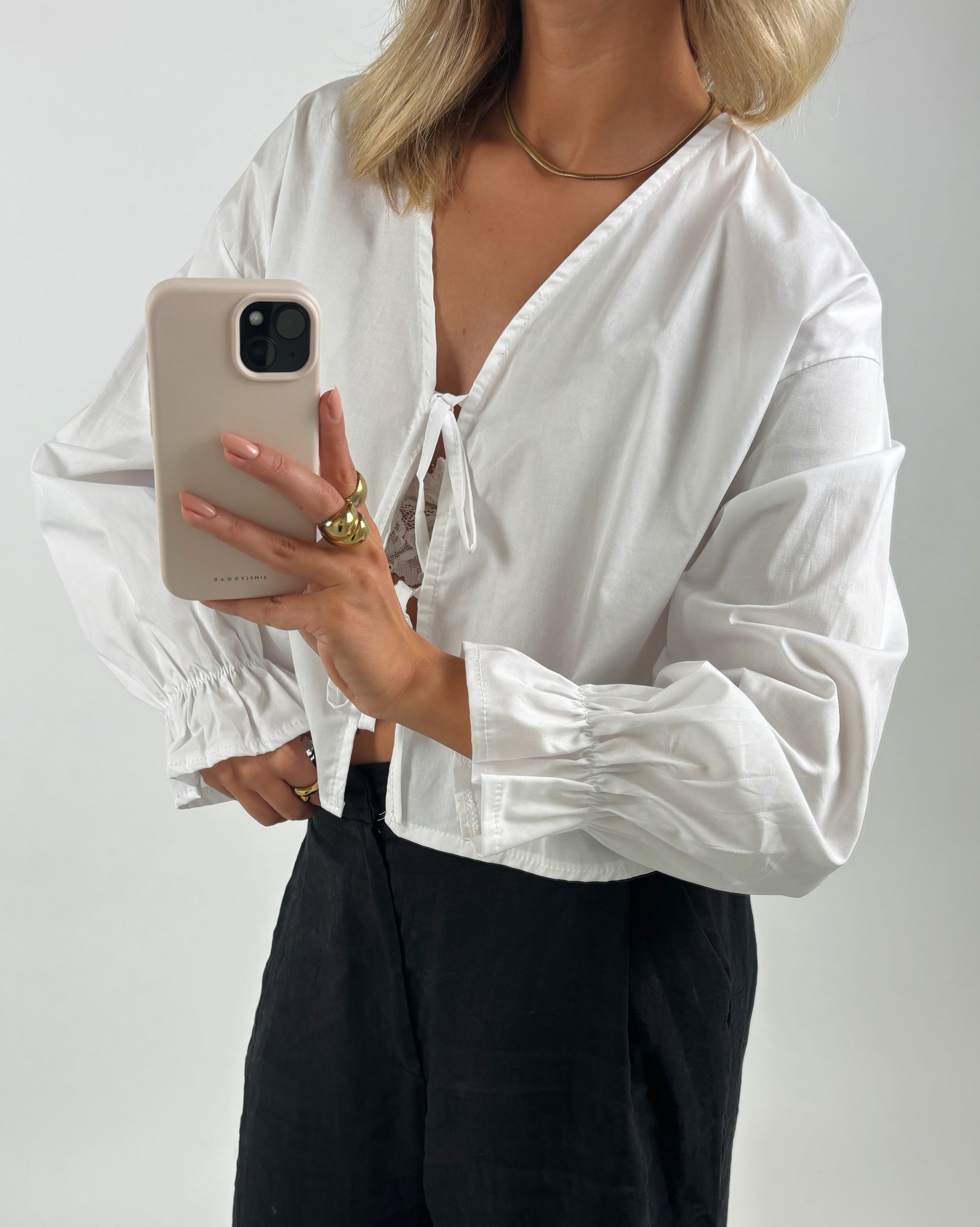 En person iført hvid skjorte tager en selfie med en mobiltelefon med beige cover. De har guldsmykker på og står foran en neutral baggrund.