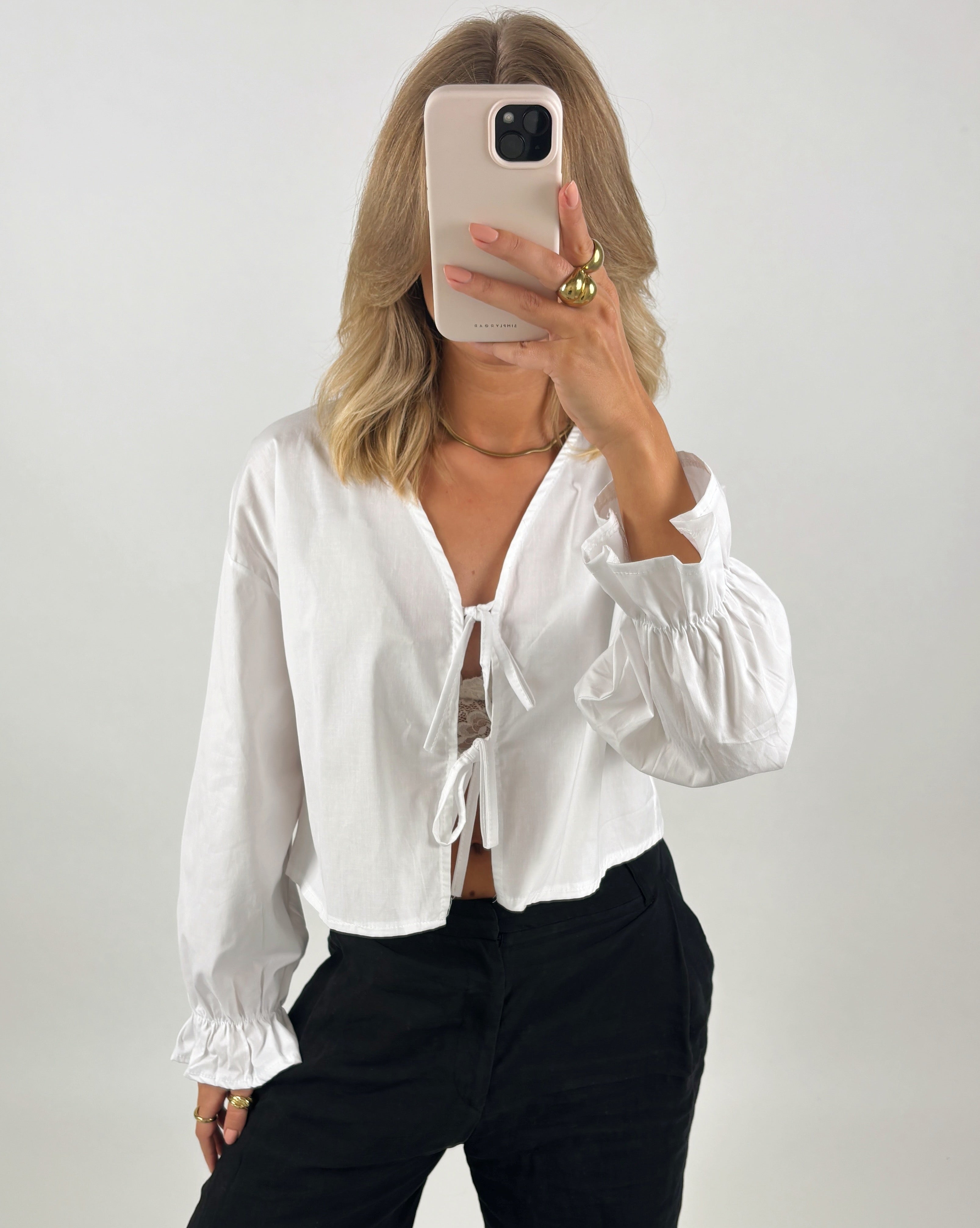 En person iført en hvid bluse holder en smartphone foran ansigtet. Blusen har bindebånd på forsiden, og personen har gyldne smykker. Baggrunden er ensfarvet og lys.