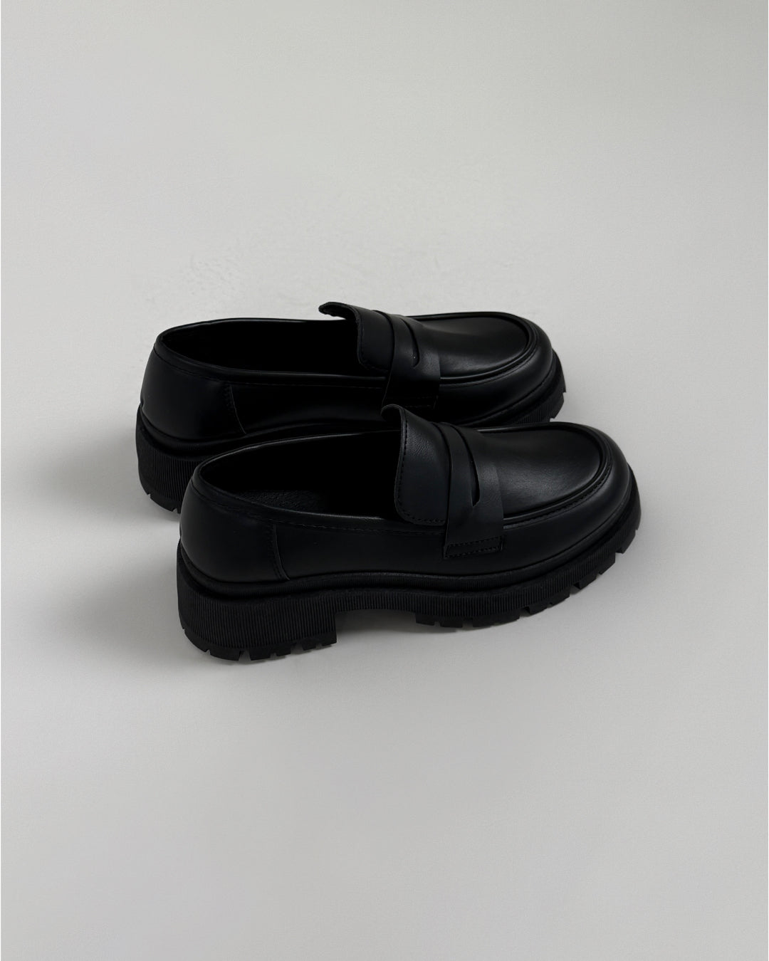 Et par sorte loafers står side om side på en lys baggrund. Skoene har en robust, rillet sål og en glat overflade med en strop over forfoden.