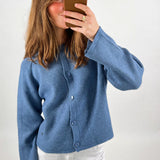 En person iklædt en blå cardigan tager et spejlbillede med en smartphone i brun cover. Personen står i en simpel, lys baggrund.