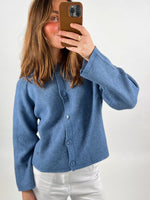 En person iklædt en blå cardigan tager et spejlbillede med en smartphone i brun cover. Personen står i en simpel, lys baggrund.