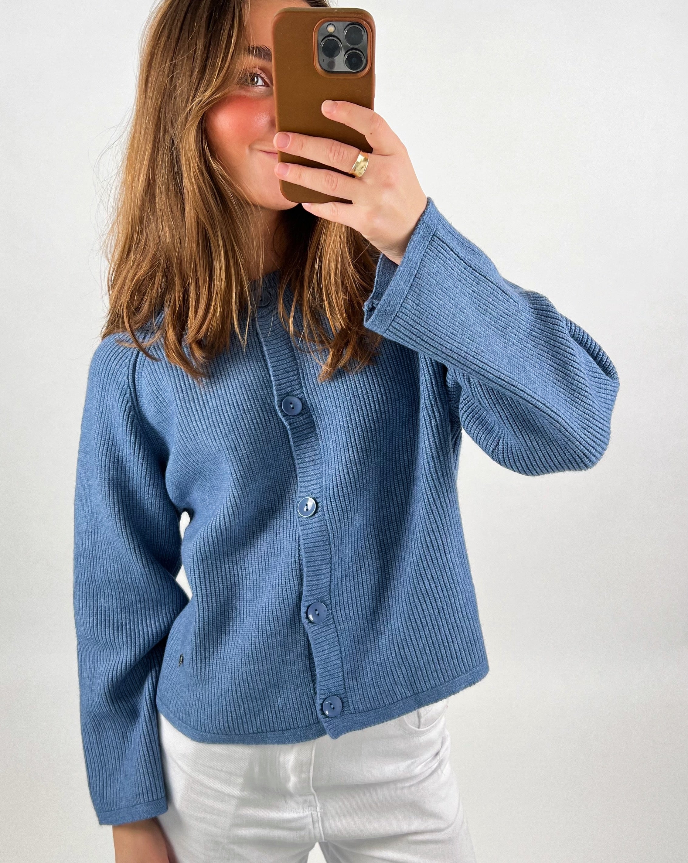 En person iklædt en blå cardigan tager et spejlbillede med en smartphone i brun cover. Personen står i en simpel, lys baggrund.