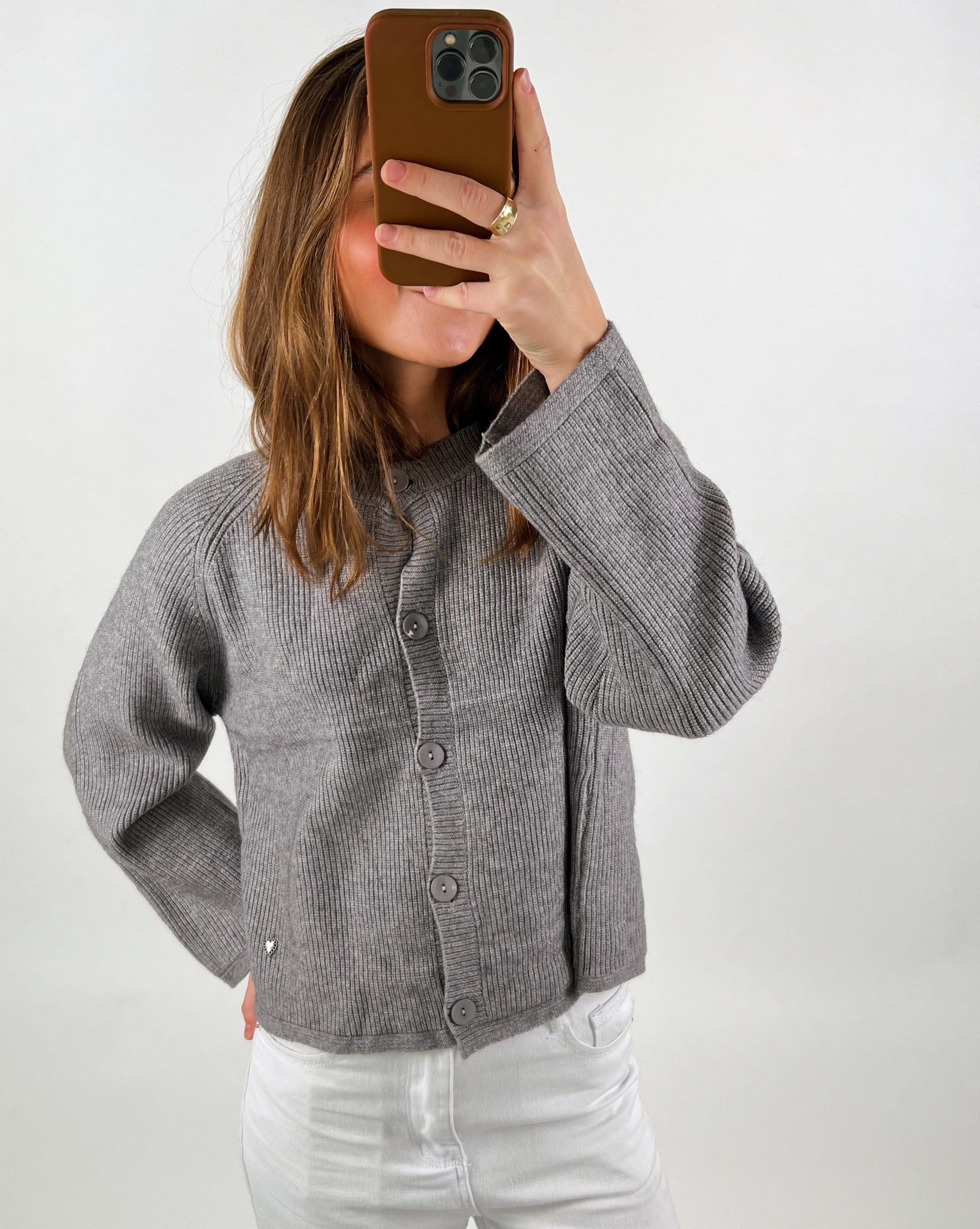 Person tager selfie i spejl, med gråt striktrøje og hvid bukser, mod neutral baggrund, bruger brun mobiltelefon som dækker ansigtet, bærer ring på fingeren.