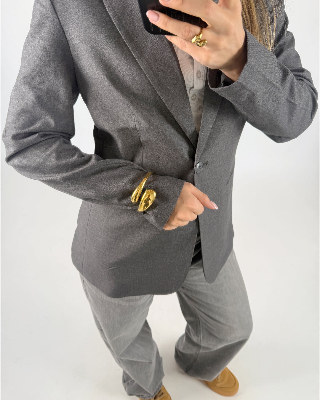 En person iført en grå blazer tager et spejl-selfie. Personen bærer store guldringe over ærmet og har grå bukser og brune sko i en lys baggrund.