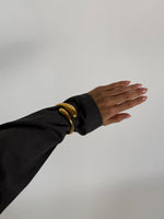 En hånd iført en mørk ærme strækker sig ud. En skinnende guldring bæres på håndleddet. Baggrunden er ensfarvet og lys, hvilket fremhæver smykket.